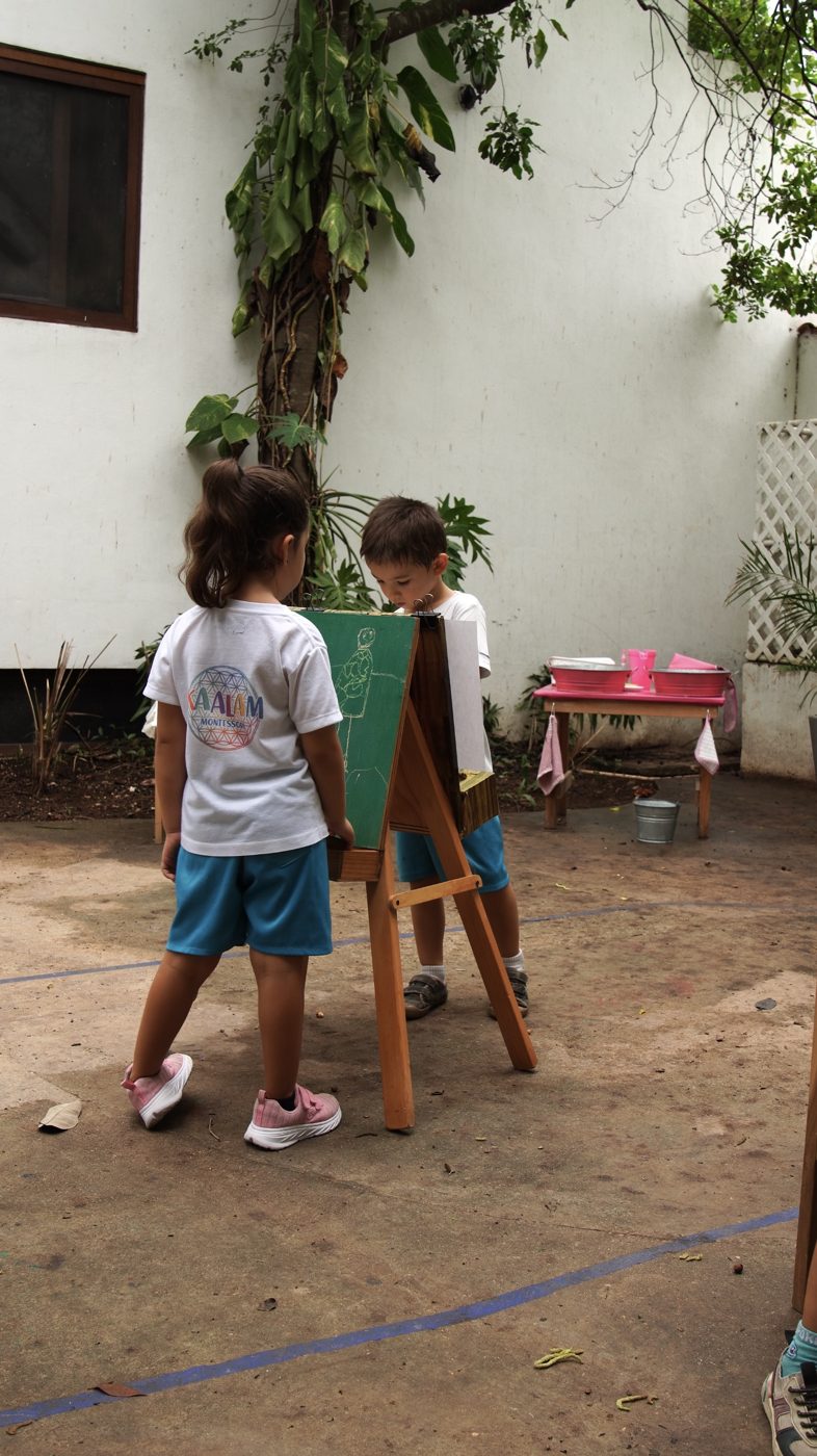 Dos niños dibujan en un caballete. La playera de la niña dice Kaalam.